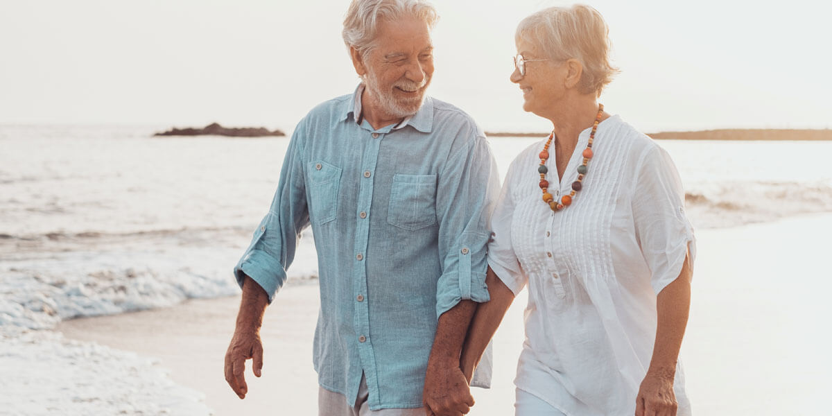 An older man and lady holding hands walking on the beach