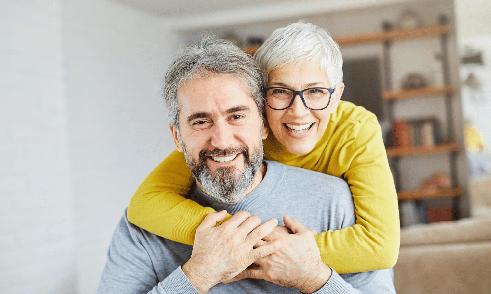 A mature women with white hair and glasses with arms around a mature man with grey hair both looking and smiling at the camera