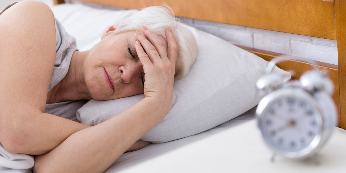 A women with white hair sleeping on white sheets with hand on head next to a blurred out alarm clock